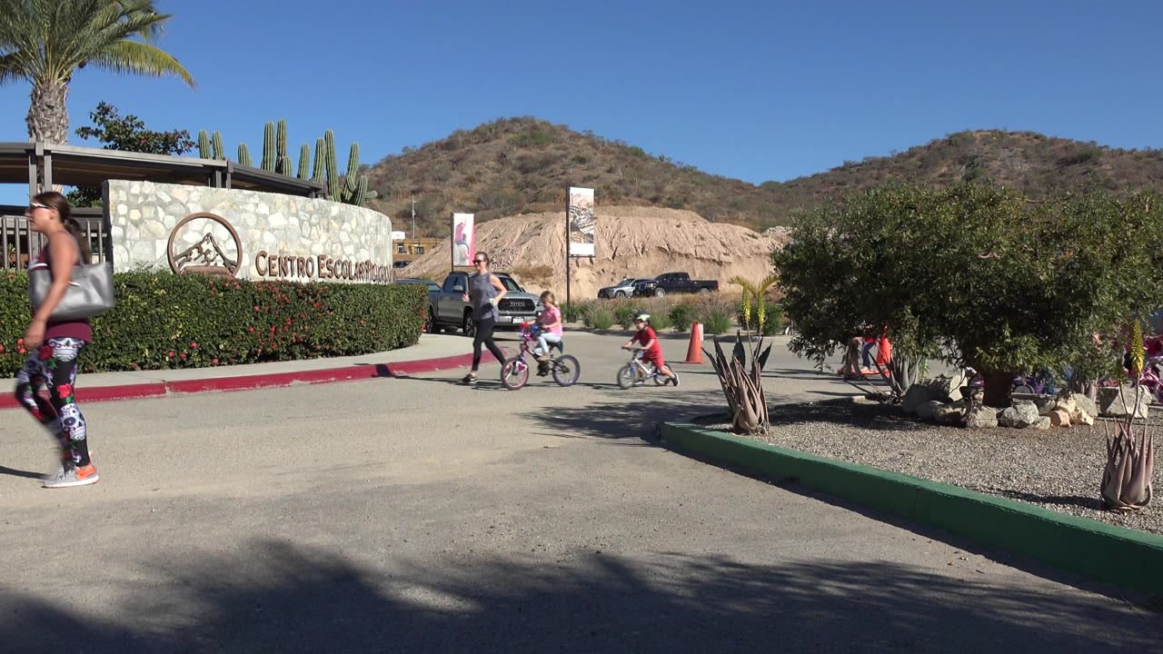 At the start of the clip in Los Cabos, Mexico, adults and children are visible near a stone visitor center sign, with bikes, scooters, hedges, desert plants, and hills behind them.