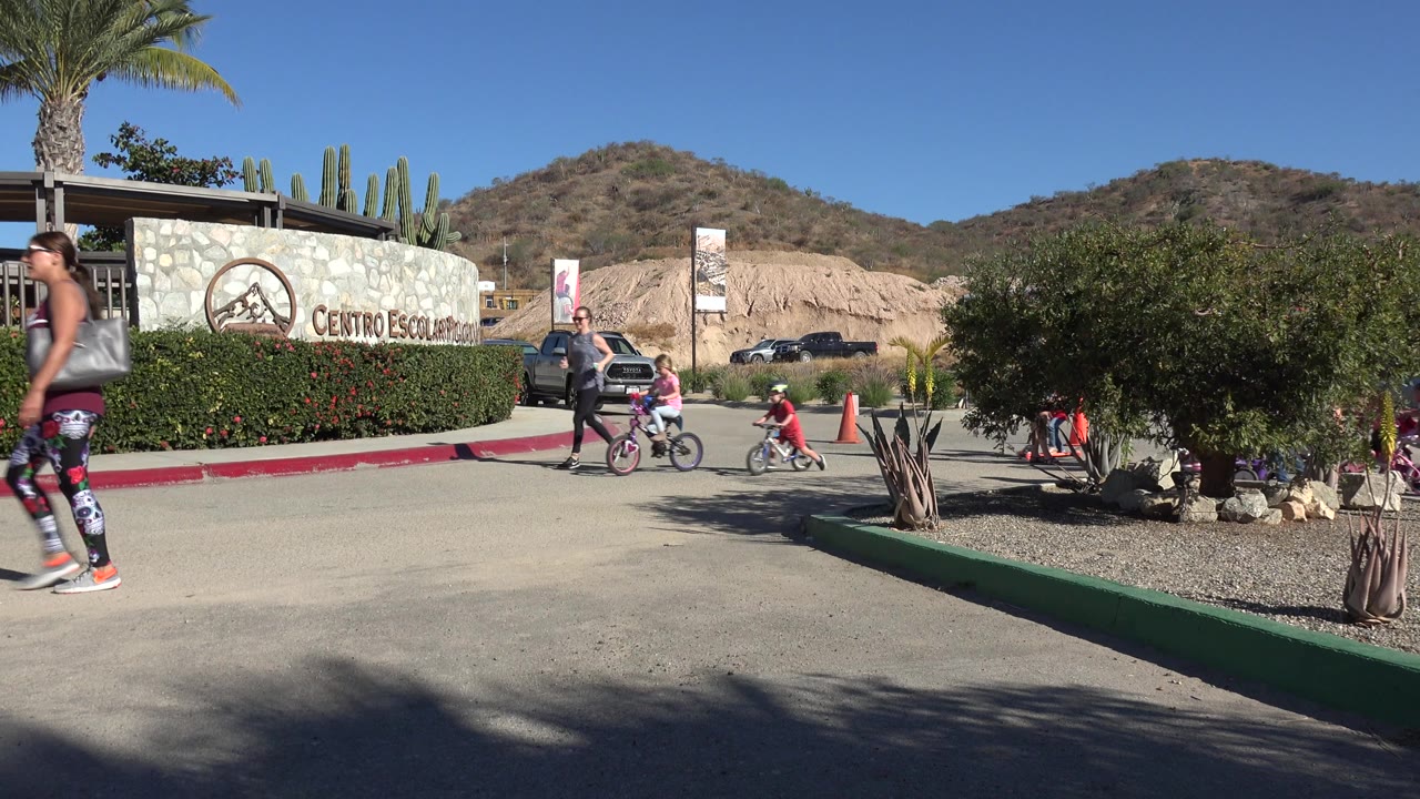 In Los Cabos, Mexico, a wide exterior view shows a woman walking in the foreground while children ride a pink bike and a small red scooter near the visitor center sign.