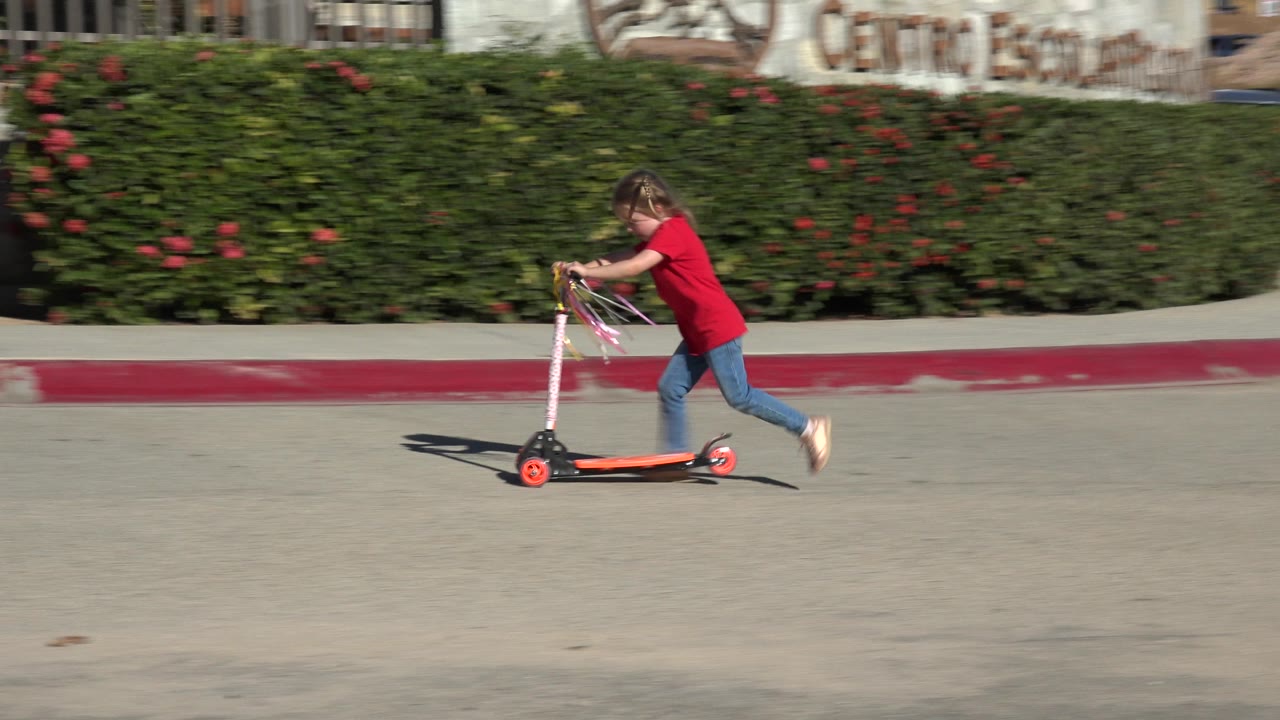 Child in a red shirt pushes a small scooter on a cement road.