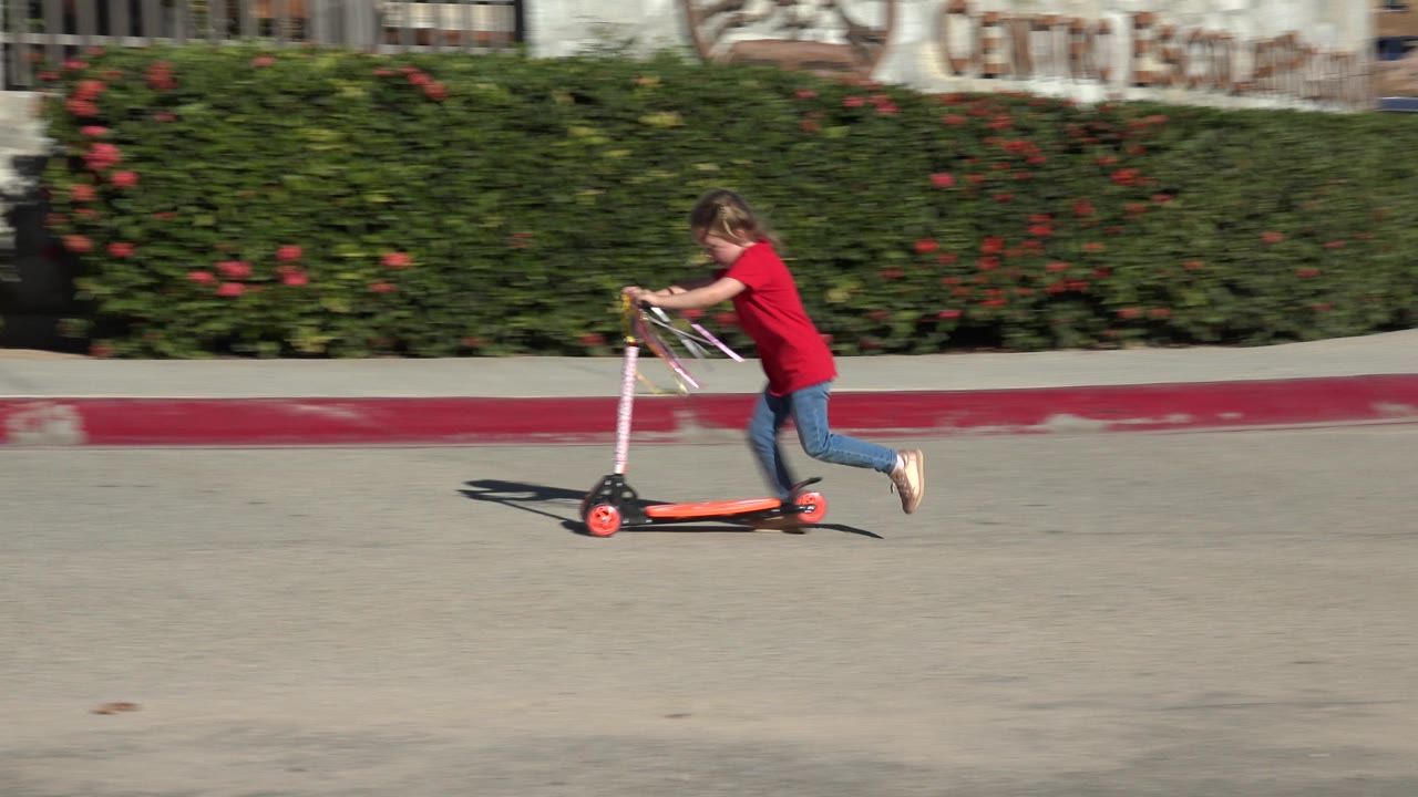 In Los Cabos, Mexico, the child on the small scooter moves farther along the cement road, still framed against hedges, a fence, and red curb paint.