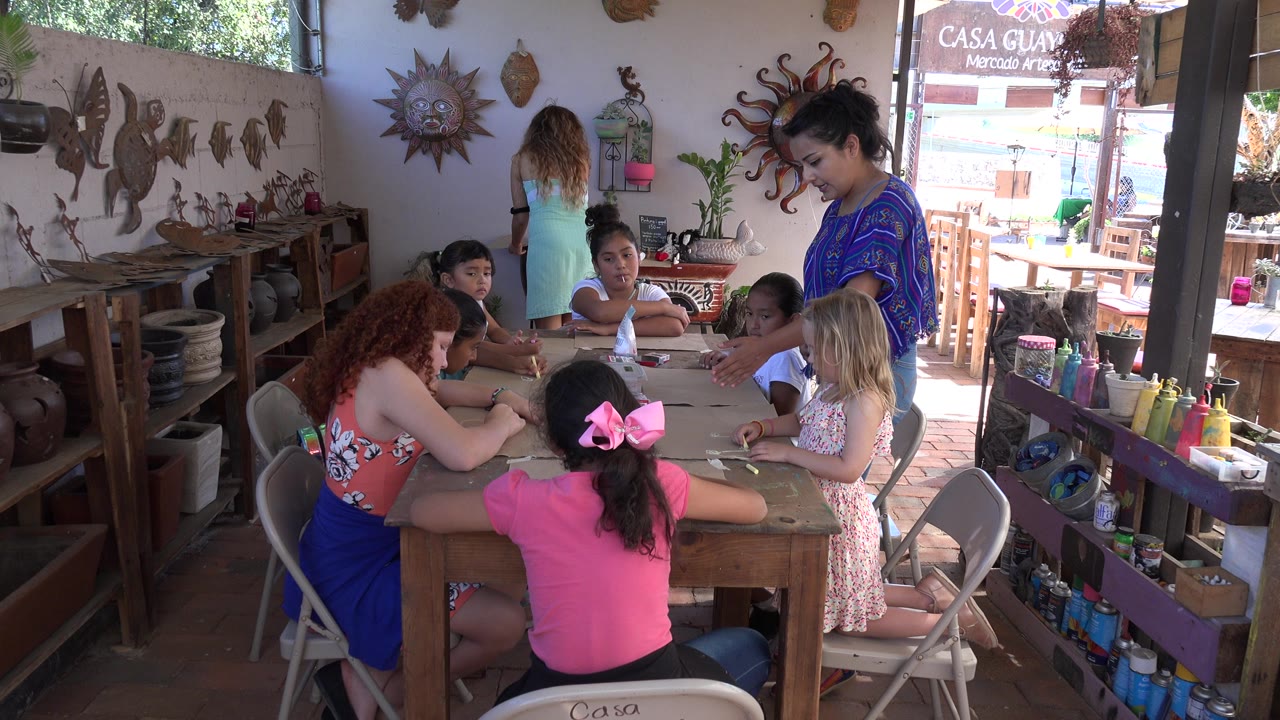 Girls work at a wooden art table while two women stand nearby.