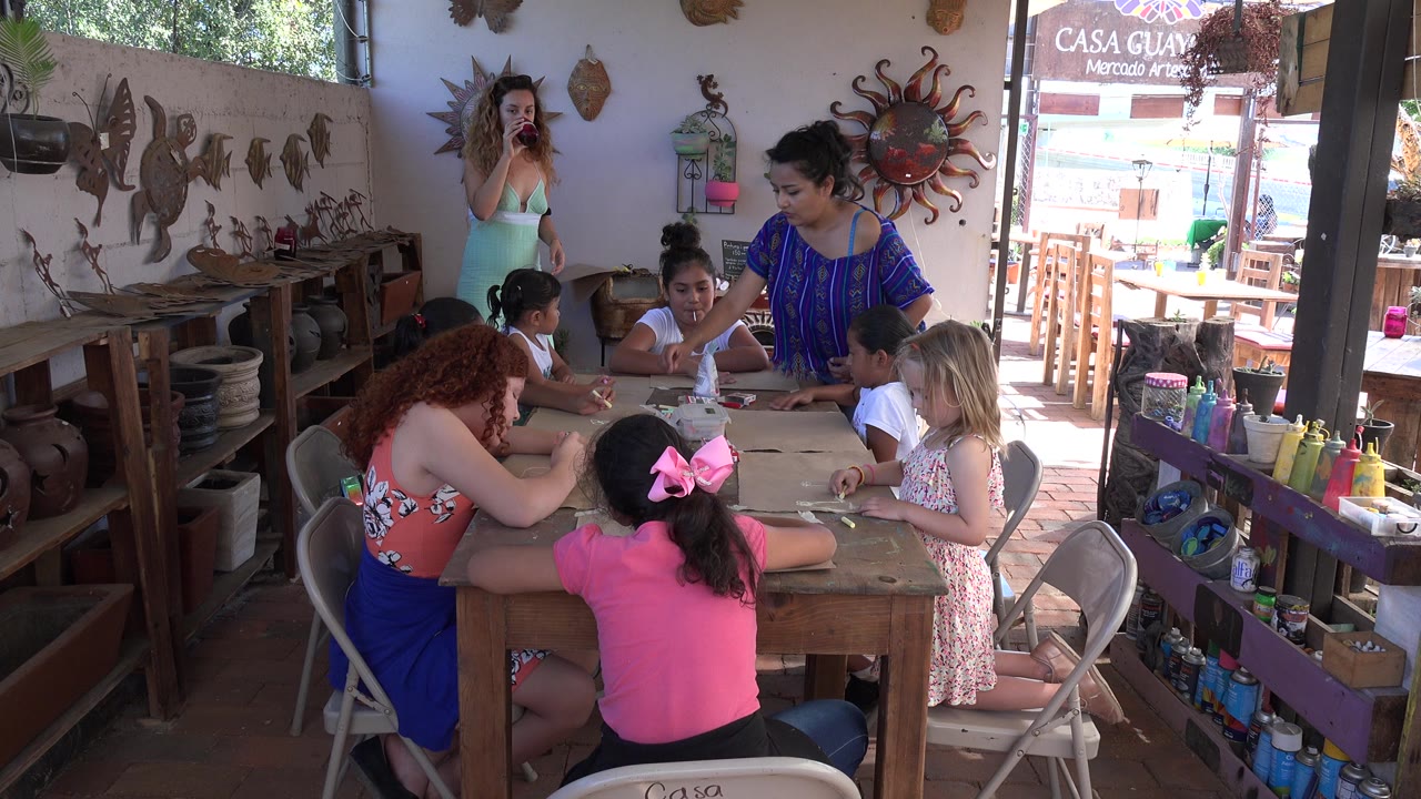 In Los Cabos, Mexico, an adult woman in a blue top stands beside children at the table while another woman stands near the back wall with craft decor around them.