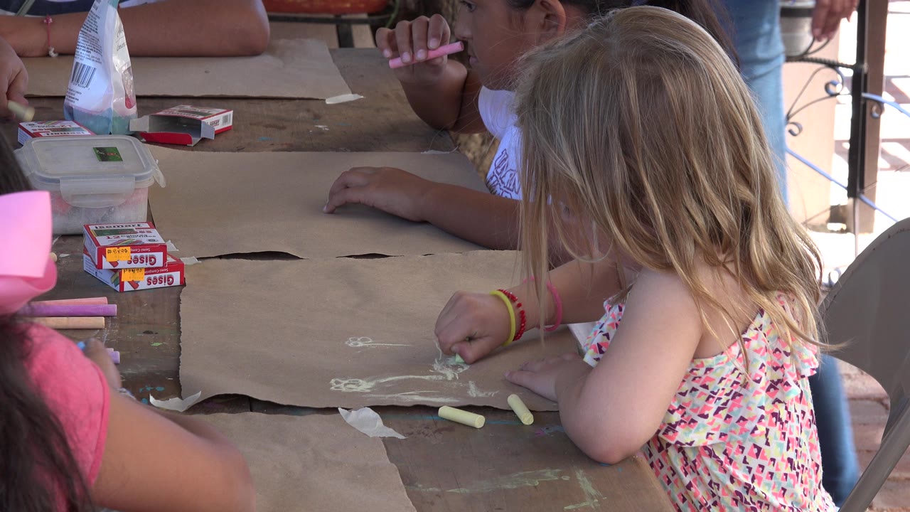 Children in Los Cabos, Mexico draw with chalk on brown paper at a shared table, with a blonde child in the foreground and other children nearby.