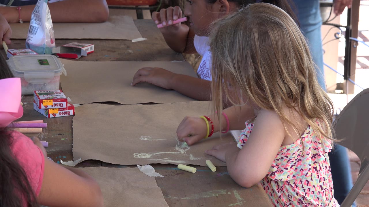 A blonde child in Los Cabos, Mexico leans over pale chalk marks while another child at the table holds a pink chalk stick.