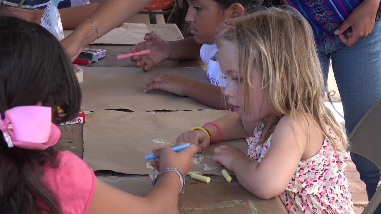 A blonde child leans over the brown paper with chalk in hand as another child with a large pink bow sits in the foreground and adults remain beside the table in Los Cabos, Mexico.