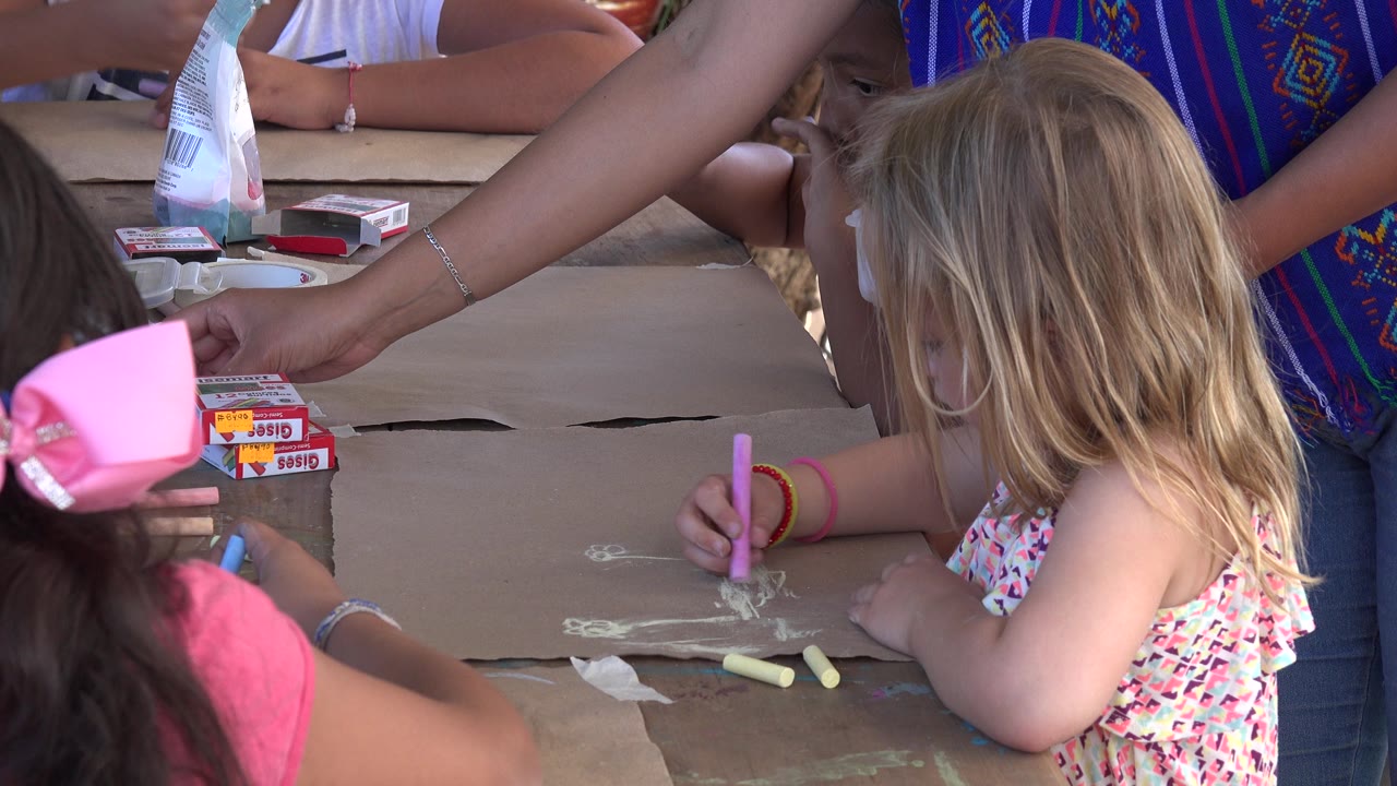 A young blonde girl in Los Cabos leans over the brown paper with a pink crayon, with another child in a pink shirt and bow partly visible in the foreground.