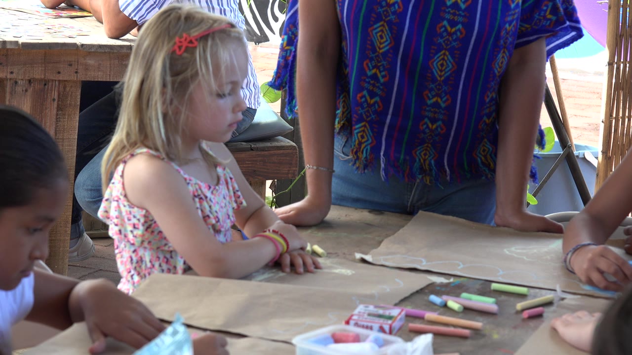 Young girl at an outdoor table with chalk and other children nearby.