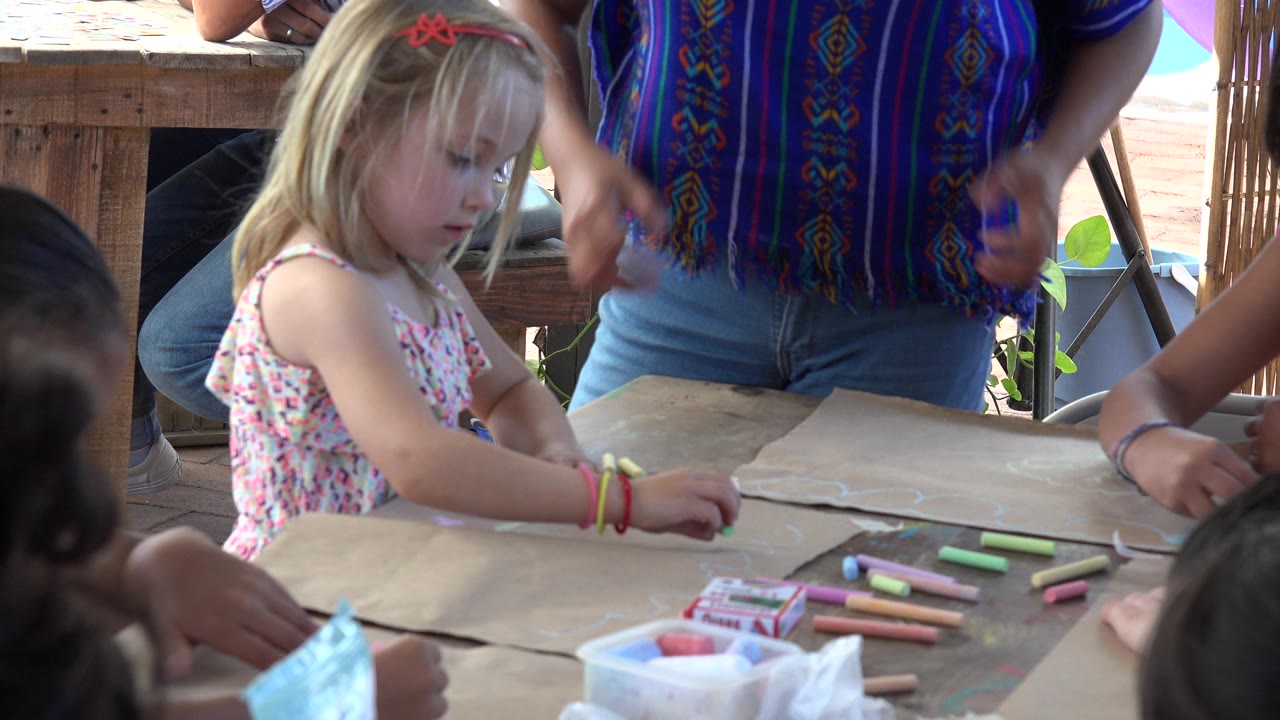 Young blonde girl drawing at a table while an adult instructor gestures nearby.