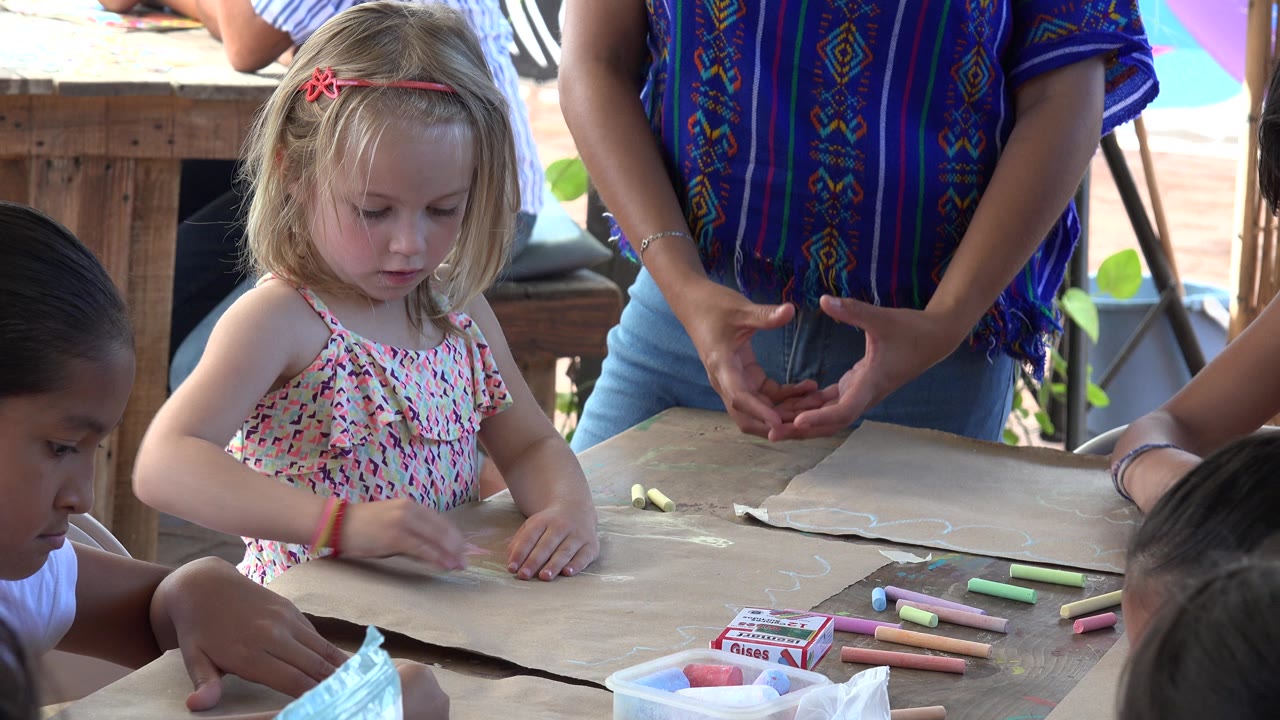 At 0:00 in Los Cabos, Mexico, a young blonde girl with a pink hairband looks down at brown paper on a wooden table while other children and an adult stand close by.