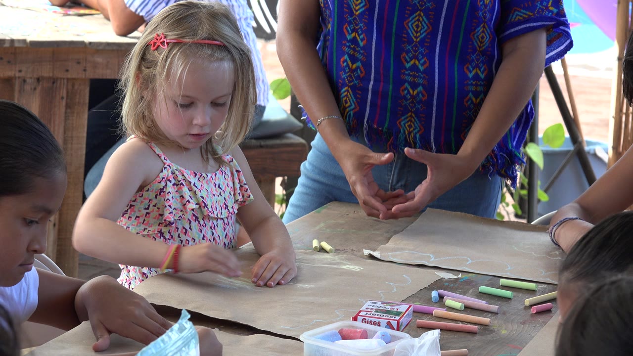 At 0:01 in Los Cabos, Mexico, the girl remains centered over the paper with chalk scattered on the table and partial faces and hands of other children at the edges.