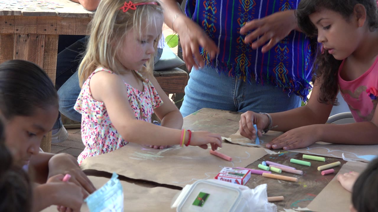 In Los Cabos, Mexico, a blonde child looks toward the table while another child in a pink shirt draws with blue chalk and the adult instructor stands behind them.