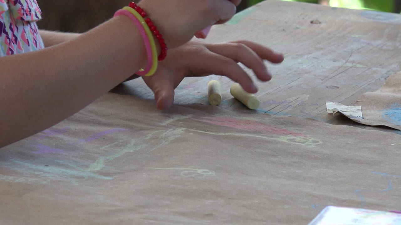 A young girl's bracelet-wearing arm reaches across the Los Cabos, Mexico tabletop toward two yellow chalk sticks on marked brown paper.