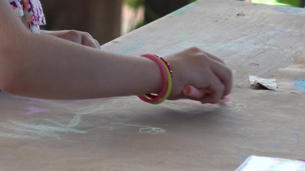 A child's forearm with colorful bracelets rests low over the chalk-covered tabletop, with faint blue and white marks visible in Los Cabos, Mexico.
