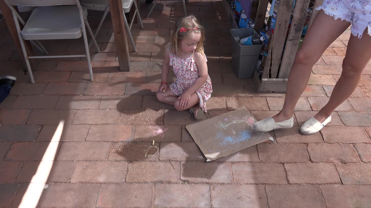 Young child sitting on patio pavers beside art paper and an adult woman's legs.
