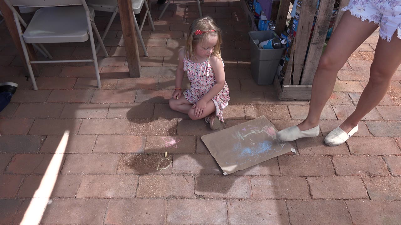 In Los Cabos, Mexico, the child remains seated on the paved patio, looking toward the art paper while the adult woman's shoes frame the right side.