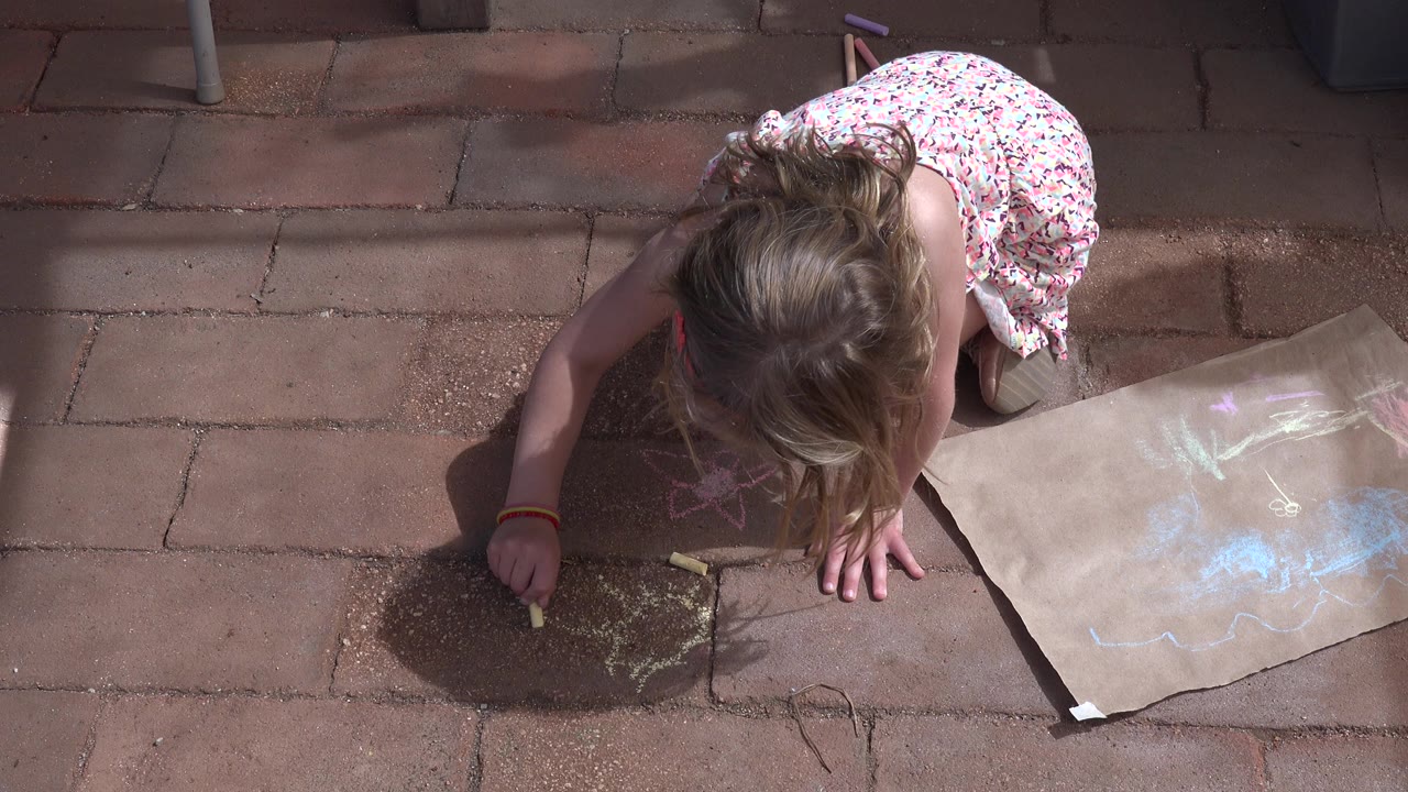 In Los Cabos, Mexico, a young girl in a floral dress leans over reddish patio pavers while drawing with yellow chalk beside a brown paper sheet with pastel marks.