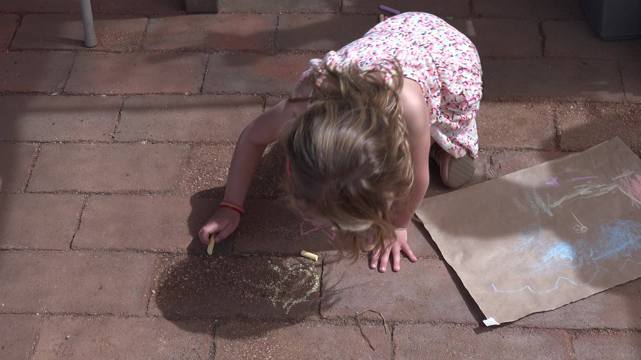 A young girl in a light patterned dress draws with chalk on reddish pavement in Los Cabos, with a partial flower shape visible near her hand and the paper shifted to the right side of frame.