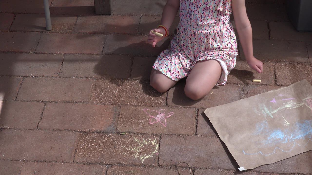 Child kneeling on patio pavers beside chalk drawings and a paper sheet.