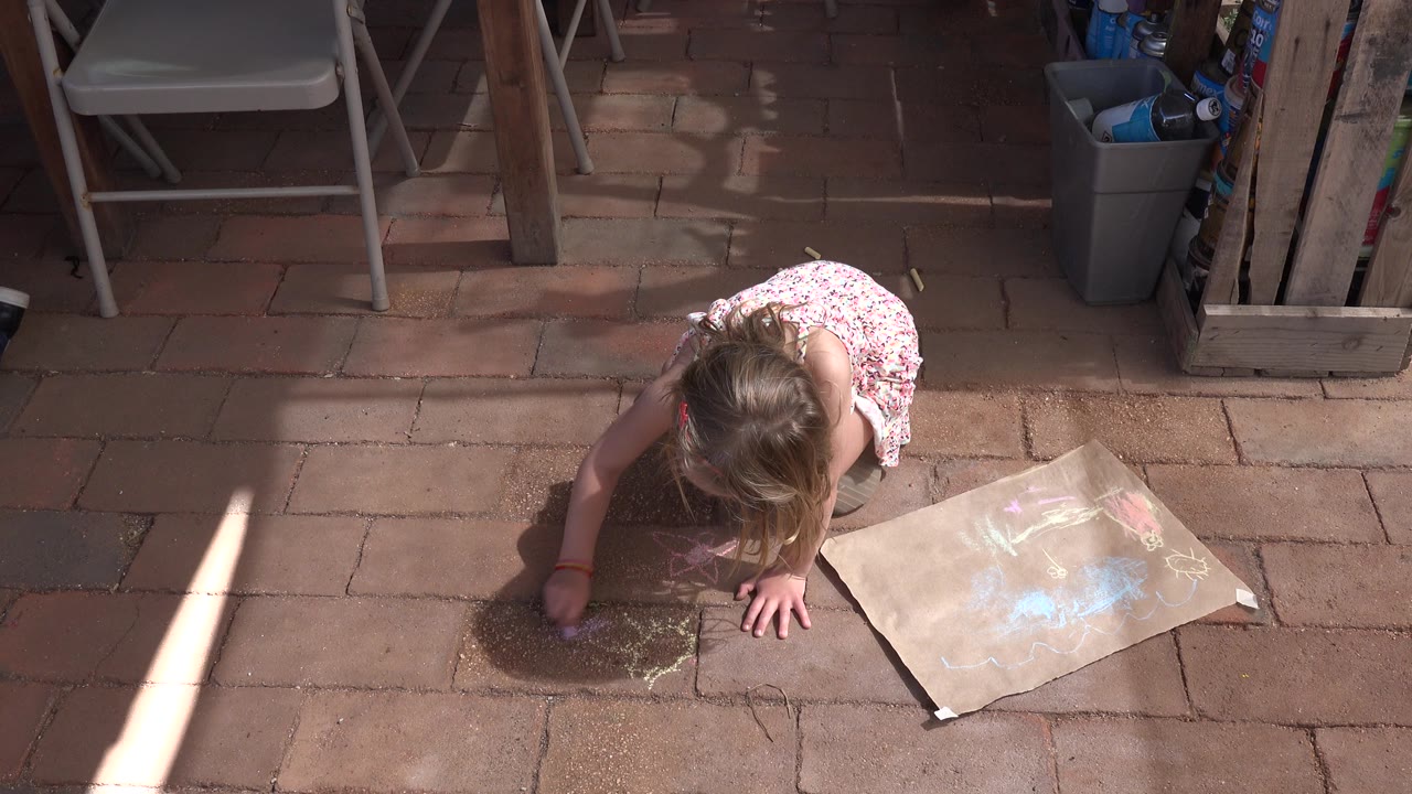 A child kneels on brick paving in Los Cabos, Mexico, drawing with sidewalk chalk beside a sheet of paper with pastel marks.