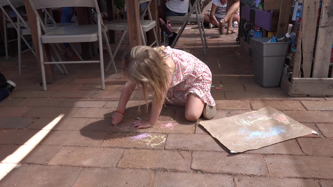 Young white girl kneels on a brick patio in Los Cabos, Mexico, starting a chalk drawing beside a cardboard sheet with patio chairs and storage items behind her.