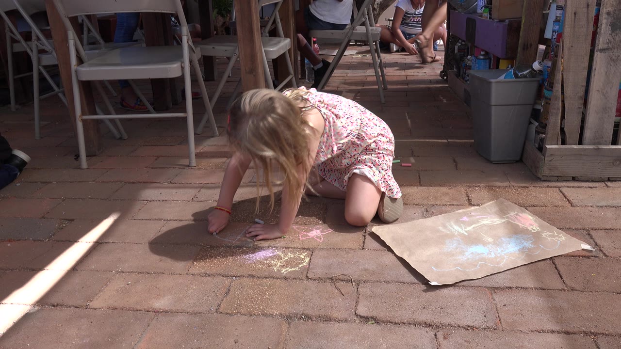 The girl stays low over the Los Cabos brick pavers, adding chalk near pink and yellow marks while the cardboard sheet lies open to her right.