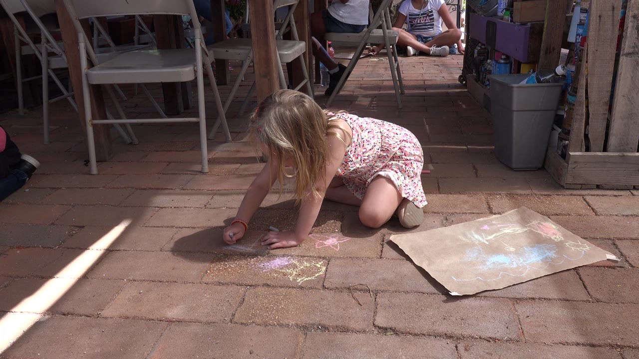 Young blonde girl kneeling on a brick patio and drawing with chalk.