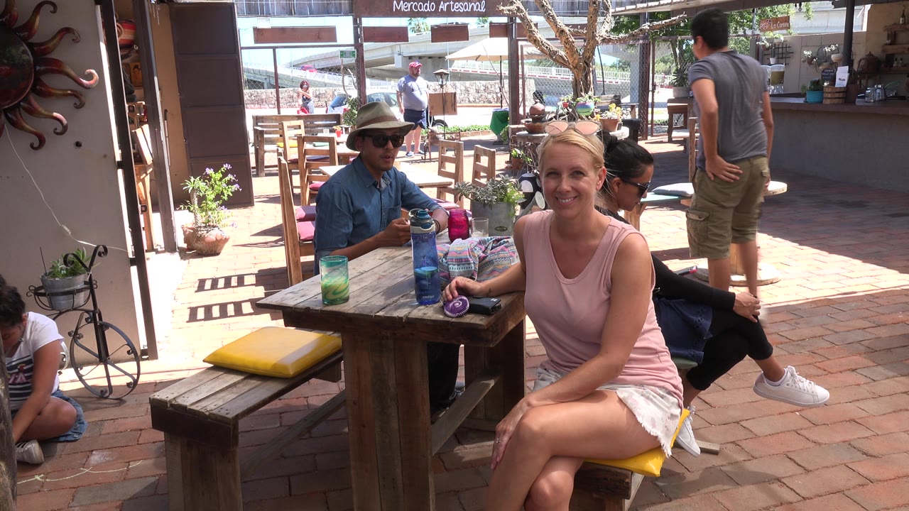 The Los Cabos cafe view holds on the smiling woman at the table while a man stands near the counter and a child sits low at the edge of the patio frame.