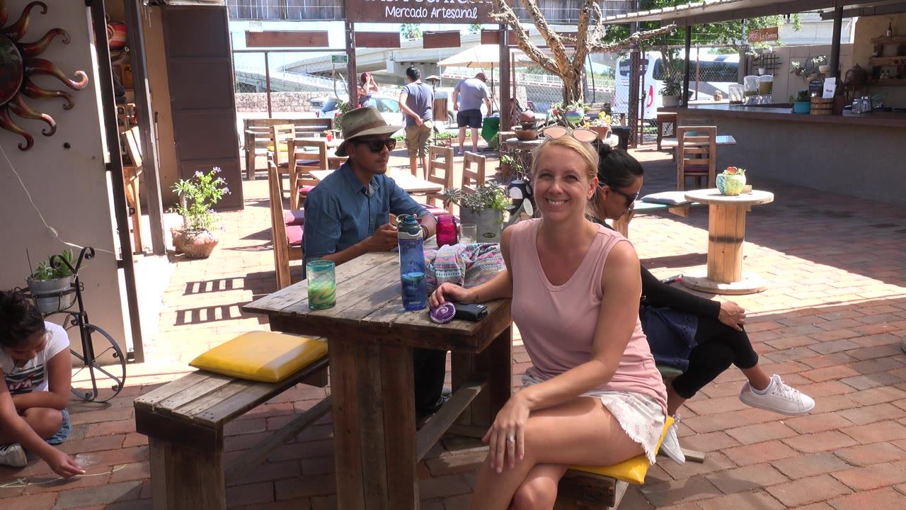 Blonde woman seated at a wooden table in a shaded patio area.