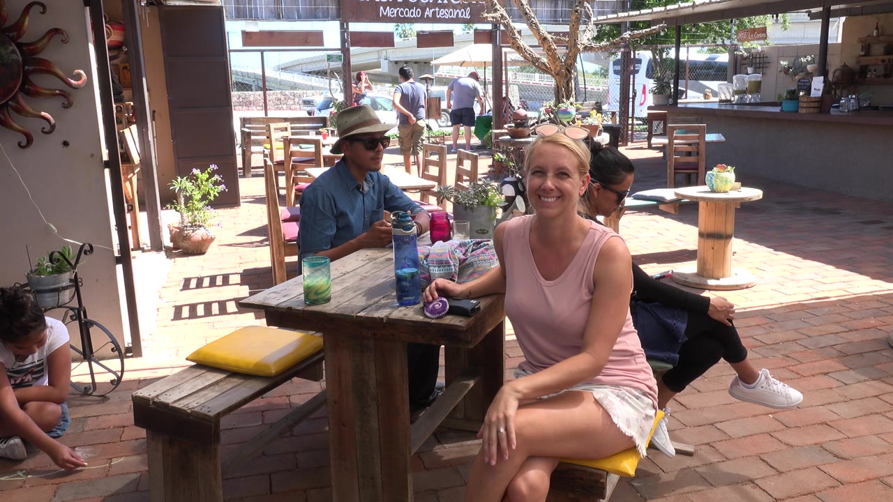 The seated woman smiles toward the videographer in Los Cabos, Mexico, while a man in a hat and another adult remain at the table behind her.