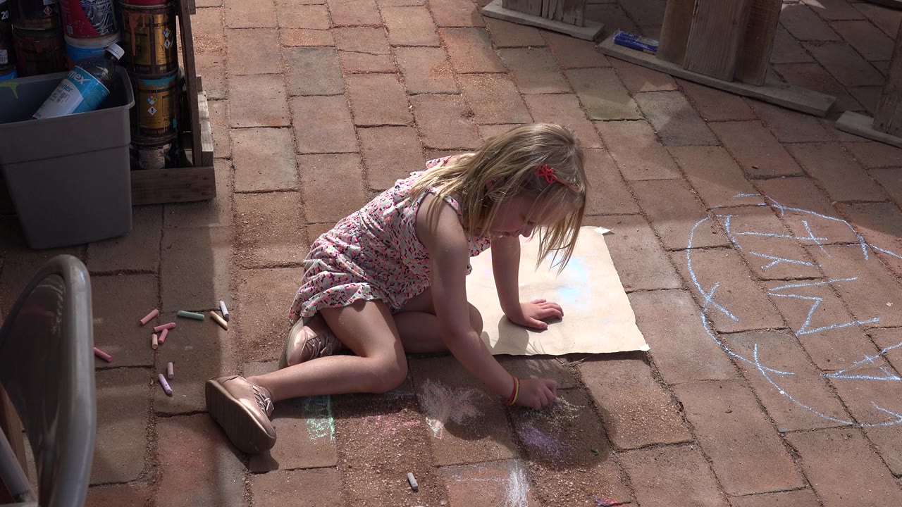 The girl leans forward over the brick paving in Los Cabos, Mexico, with her hand near a chalky patch and a paper sheet beside her knee.