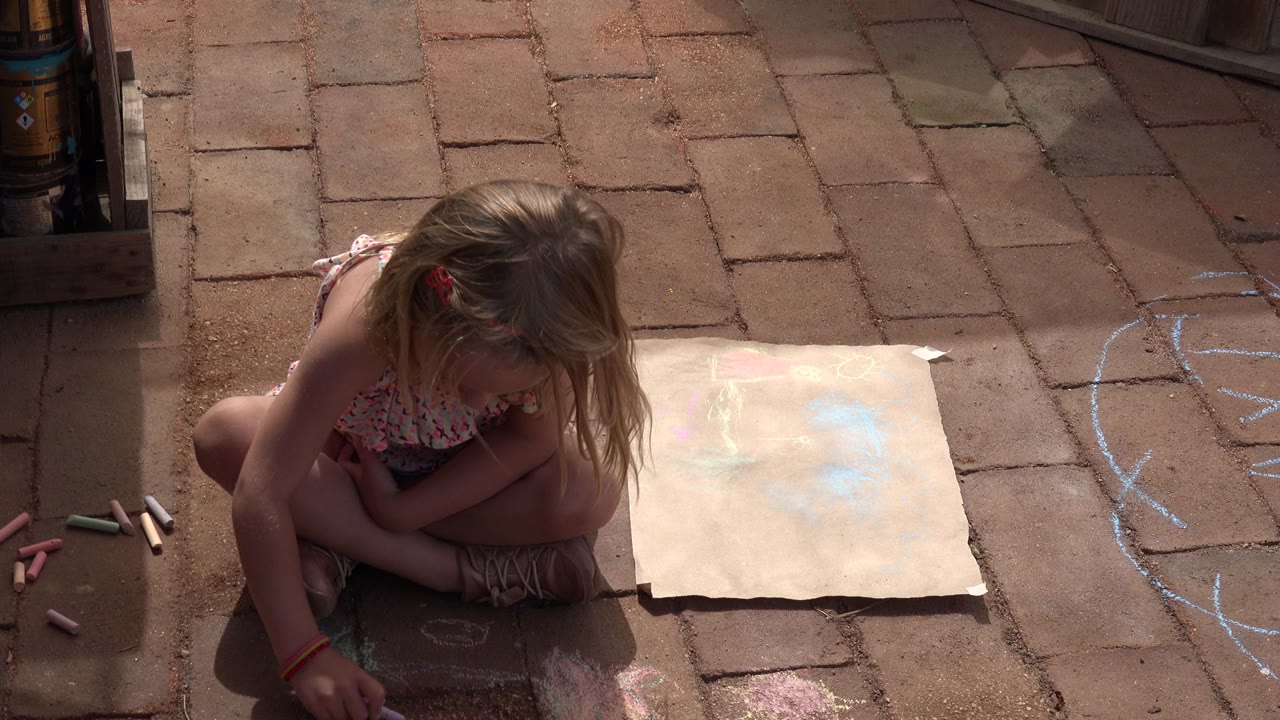 Young girl sitting on brick floor with sidewalk chalk and paper nearby.