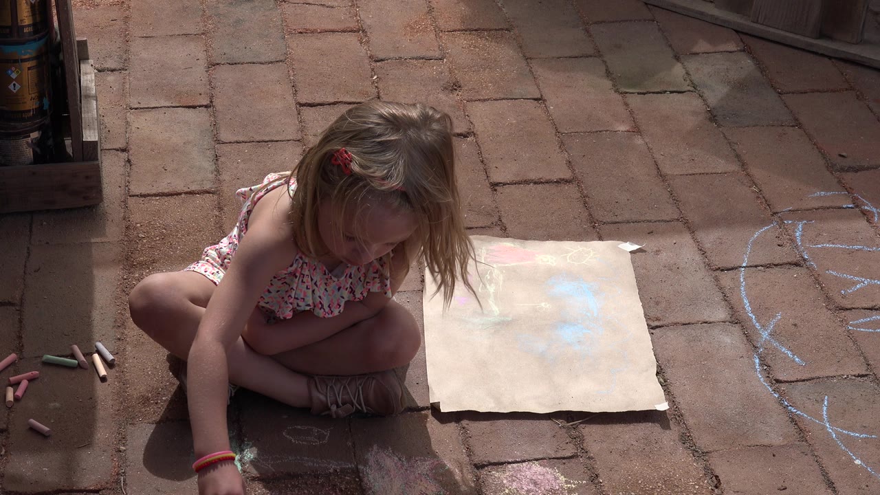 The girl leans forward on the Los Cabos brick sidewalk with chalk sticks scattered nearby and a pale drawing sheet set to her right.
