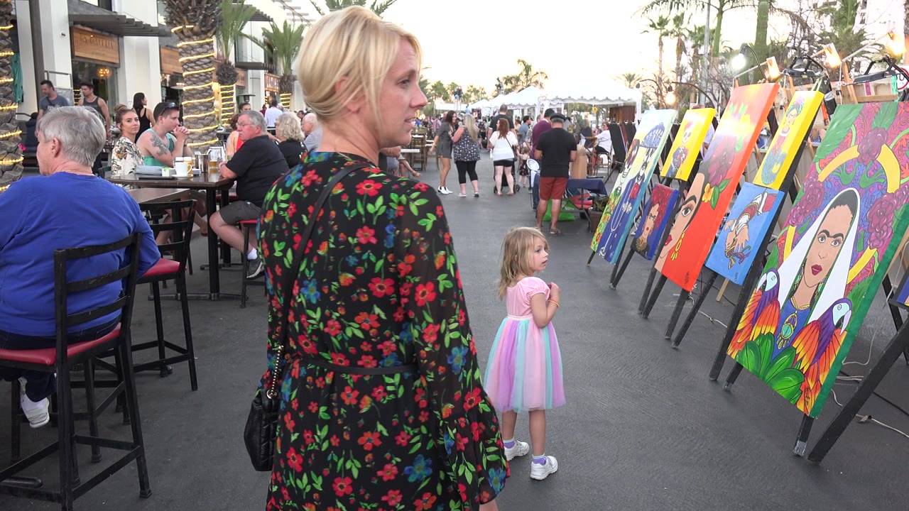 The woman and child remain beside a row of bright canvases on easels, with seated diners and pedestrians filling the Los Cabos outdoor walkway behind them.
