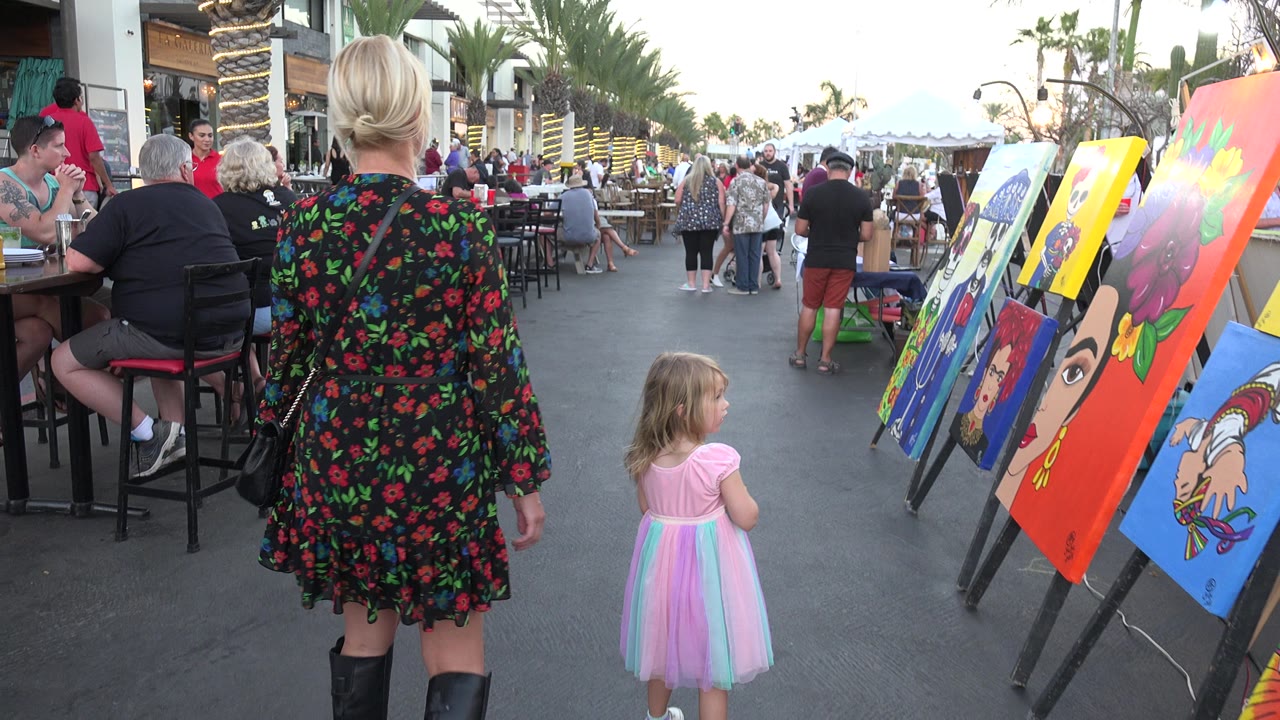 Woman and young girl walking past outdoor artwork and restaurant seating.