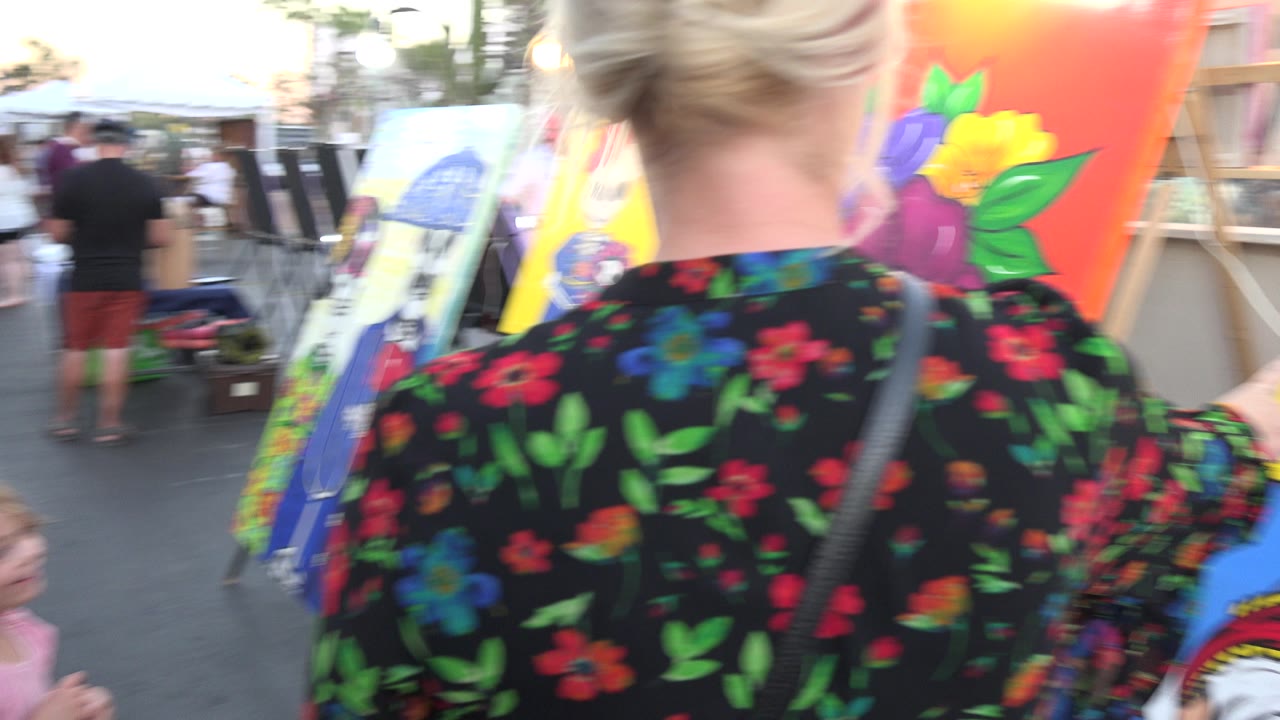 In Los Cabos, Mexico, the woman and child pass close to a row of bright canvases, including a large orange portrait with flowers and smaller illustrated panels.