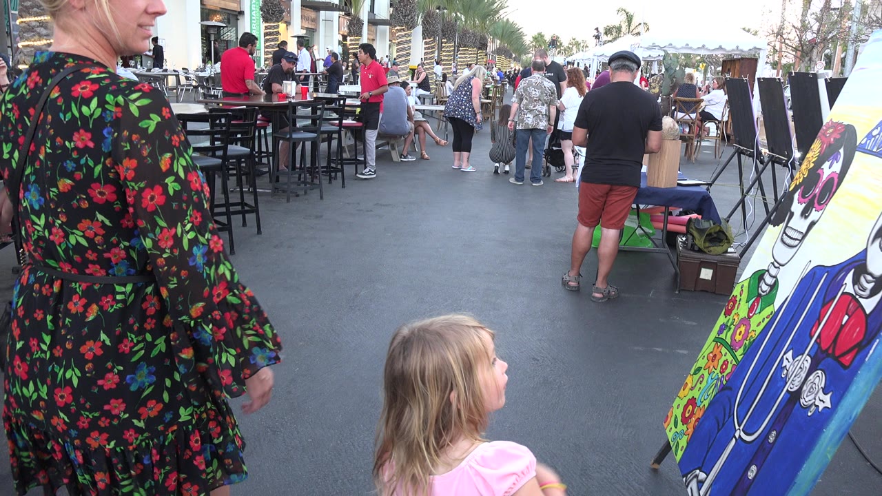 In Los Cabos, Mexico, a young girl in a pink dress walks near an adult in a black floral dress toward a blue and yellow skeleton painting beside outdoor tables.