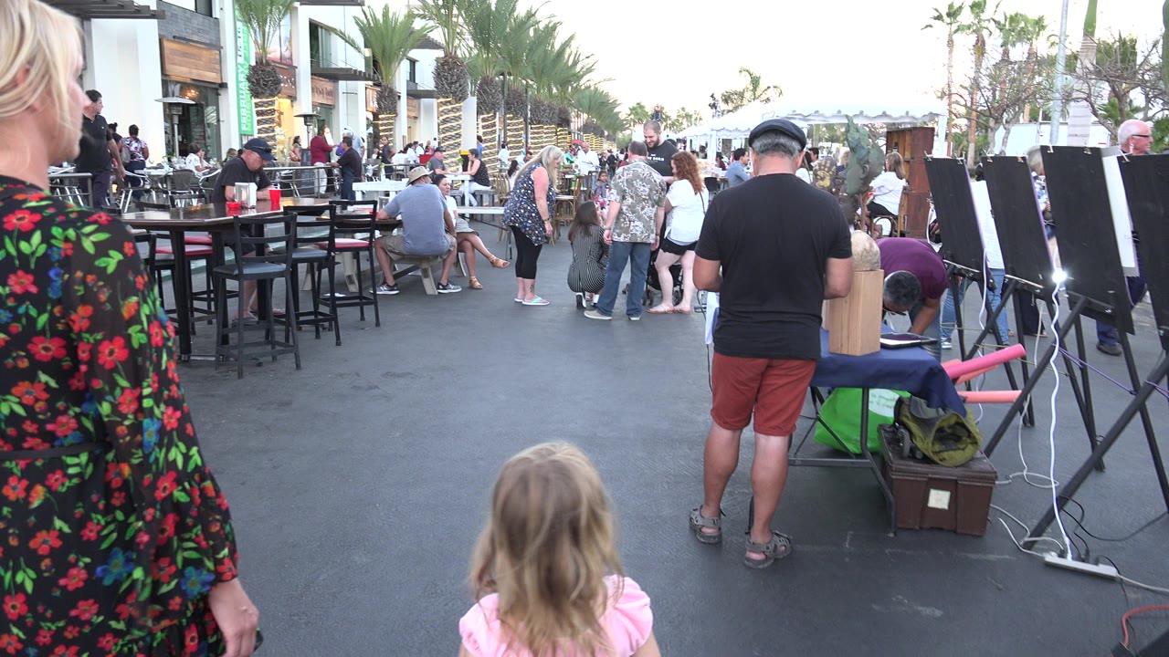 The woman and child remain in the foreground while outdoor tables, a man near display easels, storefronts, and palm-lined street activity fill the Los Cabos background.