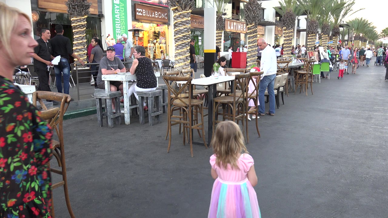 In Los Cabos, Mexico, a blonde woman in a black red-floral dress stands at the left edge while a young girl in a pink pastel dress faces down a cafe-lined walkway.
