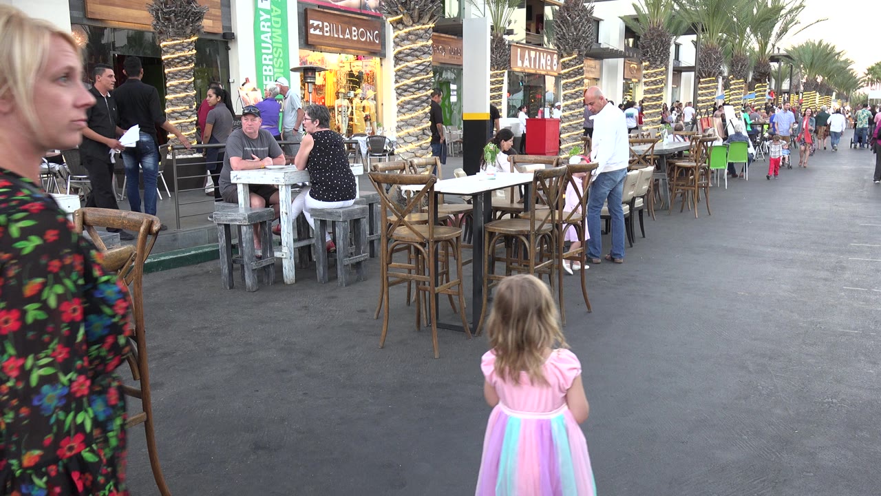 In Los Cabos, Mexico, the woman moves closer across the left foreground while the young girl remains ahead in the center of the walkway near stacked cafe chairs and storefront signs.