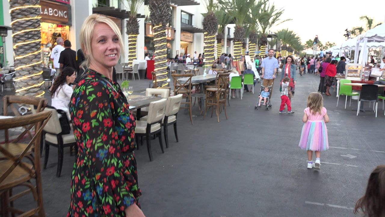 Blonde woman beside outdoor cafe seating on a busy walkway.