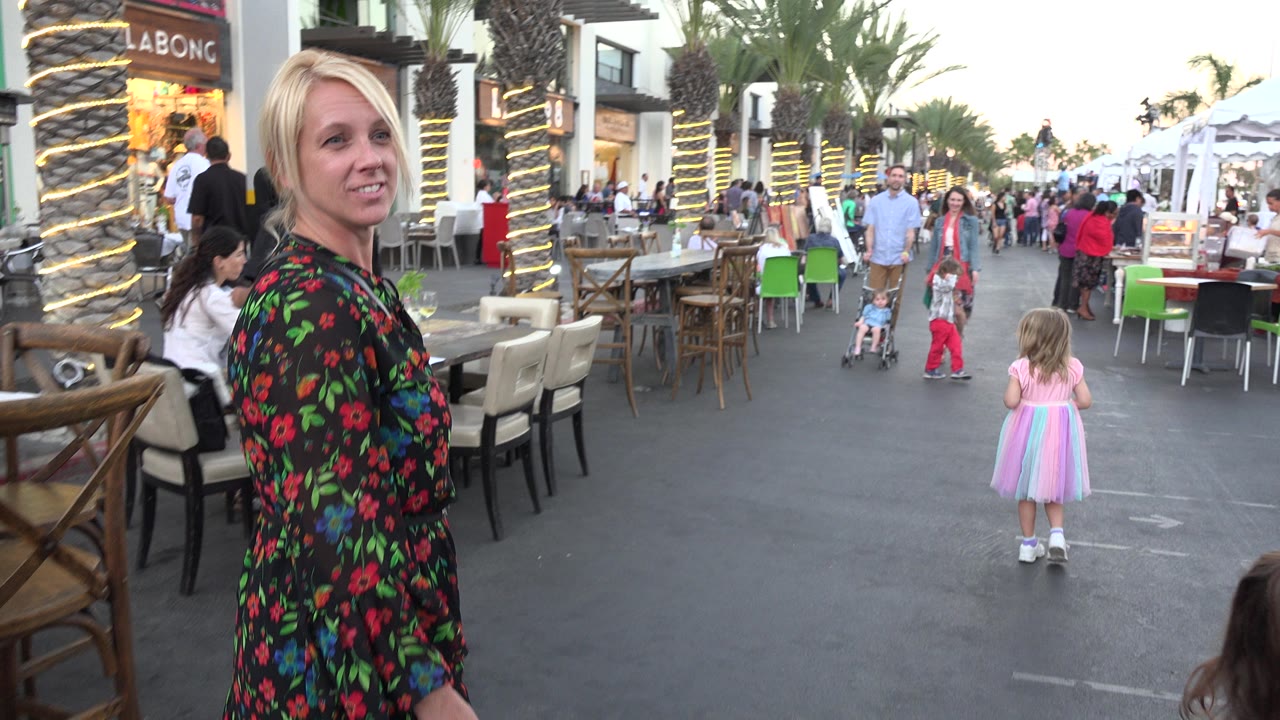 The woman stands closer to the camera on the Los Cabos walkway, with a small child in a pastel dress ahead of her and restaurant seating, palm trees, pedestrians, and shop signs behind.