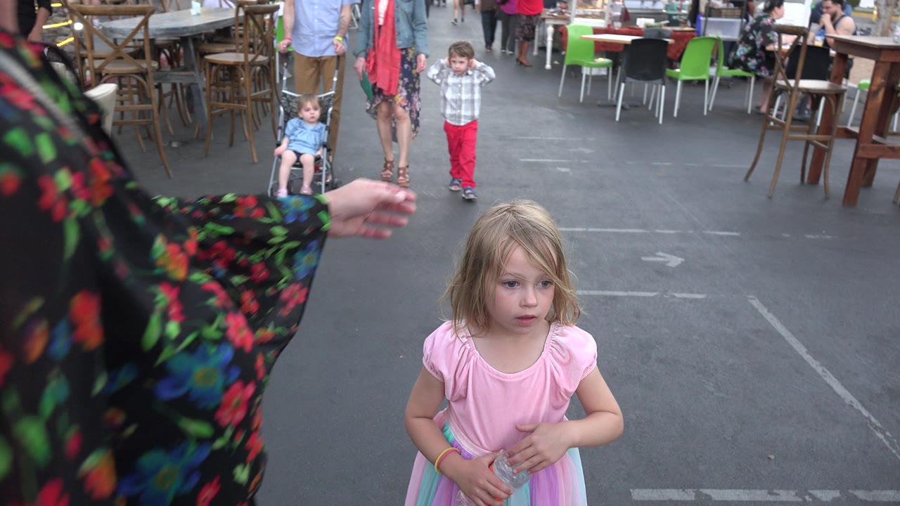 Little girl in a pink dress holding a water bottle near adults on a pedestrian street.