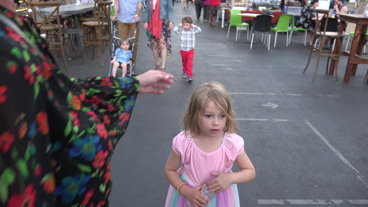 The Los Cabos street scene holds on the girl in pink as a floral sleeve fills the left foreground and a seated child, walking adults, tables, and green chairs appear behind her.