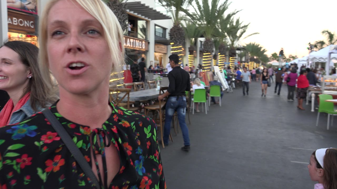 A blonde white woman in a black floral top is close to the camera in Los Cabos, Mexico, with outdoor tables, palm trees, storefronts, and several people behind her.