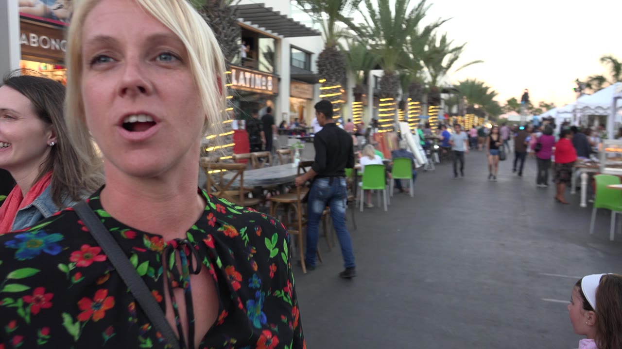 The woman faces the camera while speaking in Los Cabos, Mexico, with a second woman partly visible at the edge and pedestrians moving between outdoor seating and storefronts.