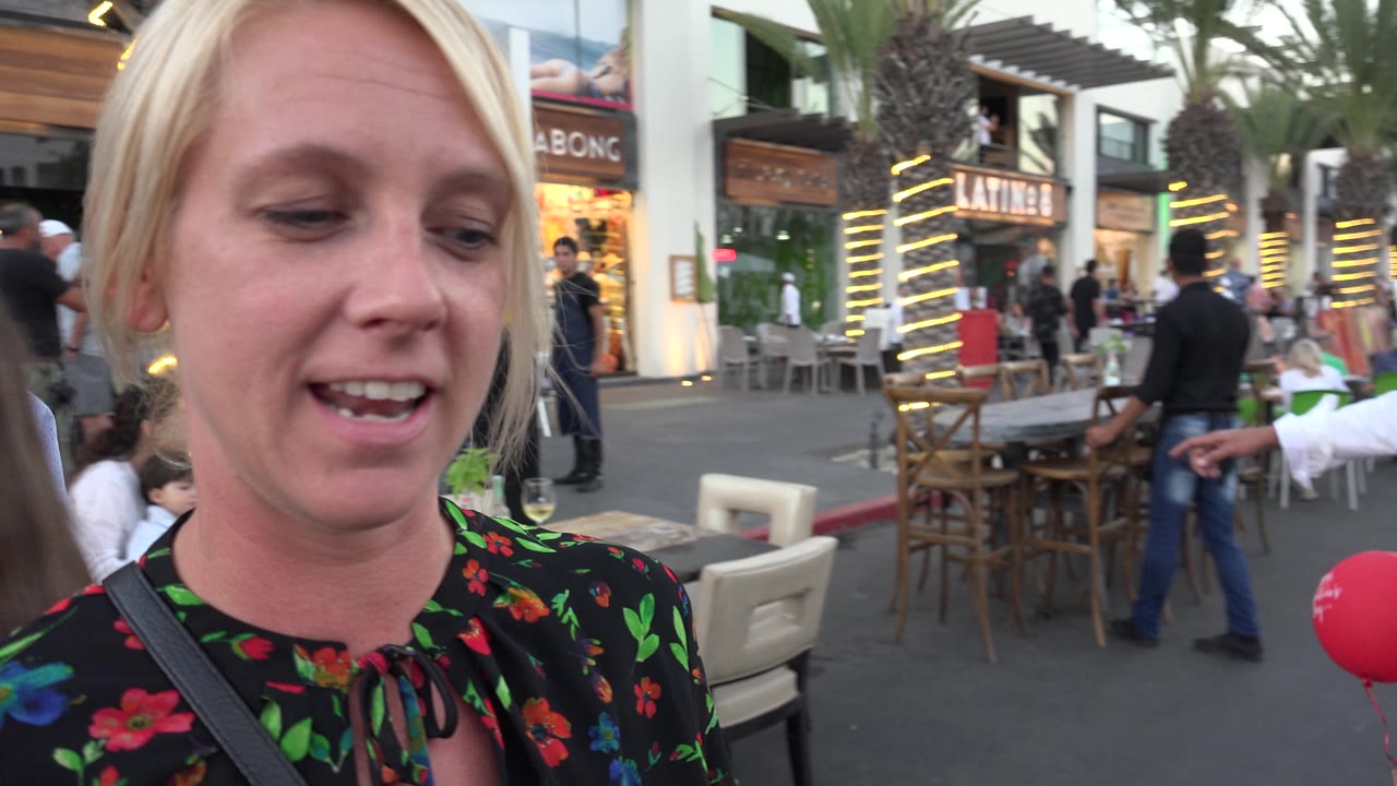 Blonde woman looks downward beside outdoor seating and storefronts.