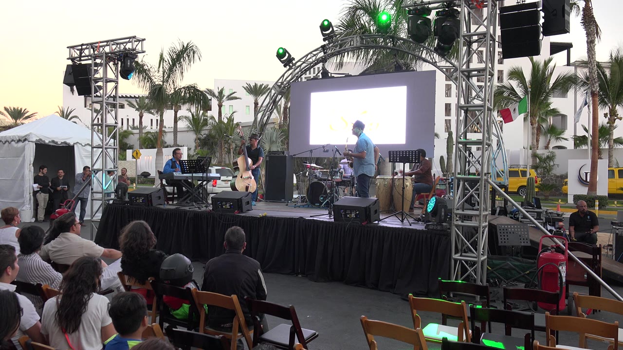 Los Cabos, Mexico outdoor event stage with keyboard player, standing string player, drums, bongos, truss lights, projection screen, and seated audience in front.