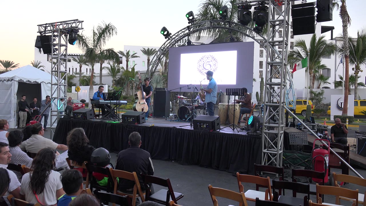 Opening frame of an outdoor band stage in Los Cabos, Mexico, with seated audience members in the foreground, truss lighting, instruments, palm trees, and a bright screen behind the performers.