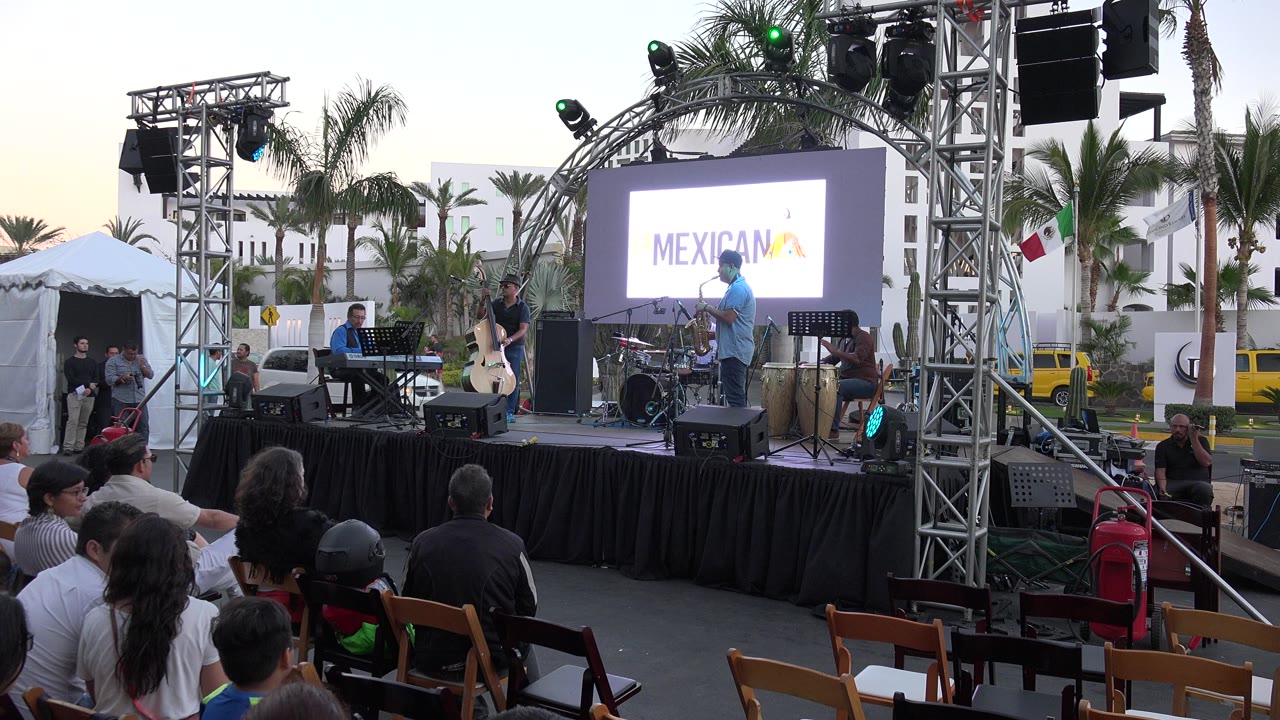 A Los Cabos outdoor stage is seen from the audience, with musicians, instruments, truss lighting, palm trees, tents, and seated people in the foreground.