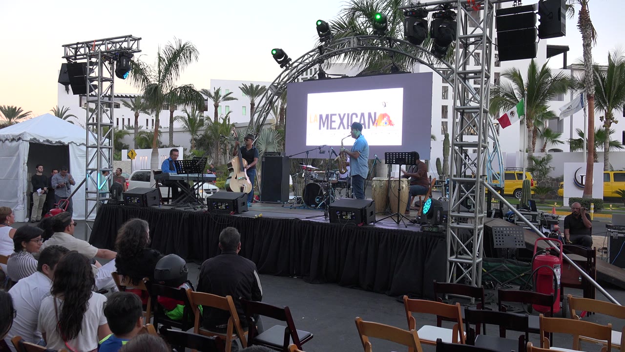 The Los Cabos stage remains centered as the LED screen shows Mexicania-style event text, with a singer at the front, a keyboardist to the left, and audience members seated below.